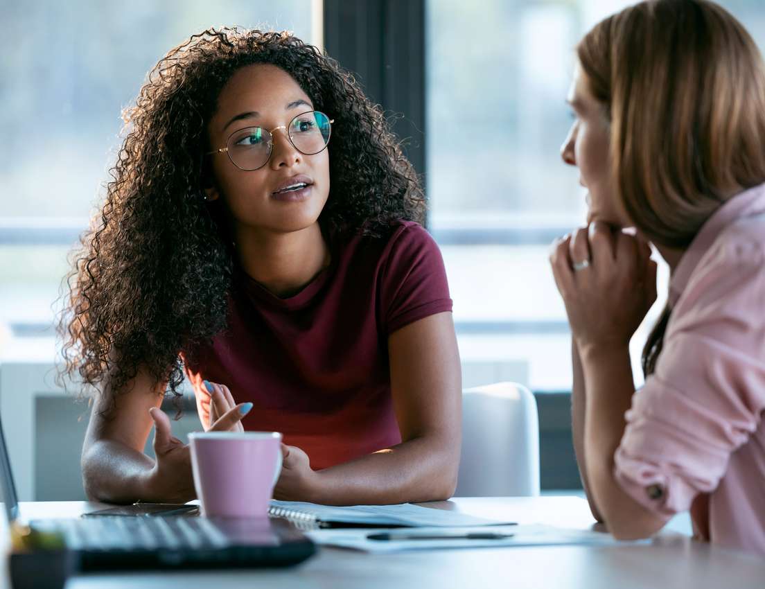 Two women in an office discussion. One with curly hair and glasses speaks, while the other listens attentively at a desk with