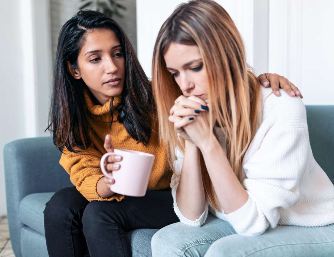 A compassionate woman in a yellow sweater comforts a distressed woman with clasped hands and bowed head on a blue sofa.