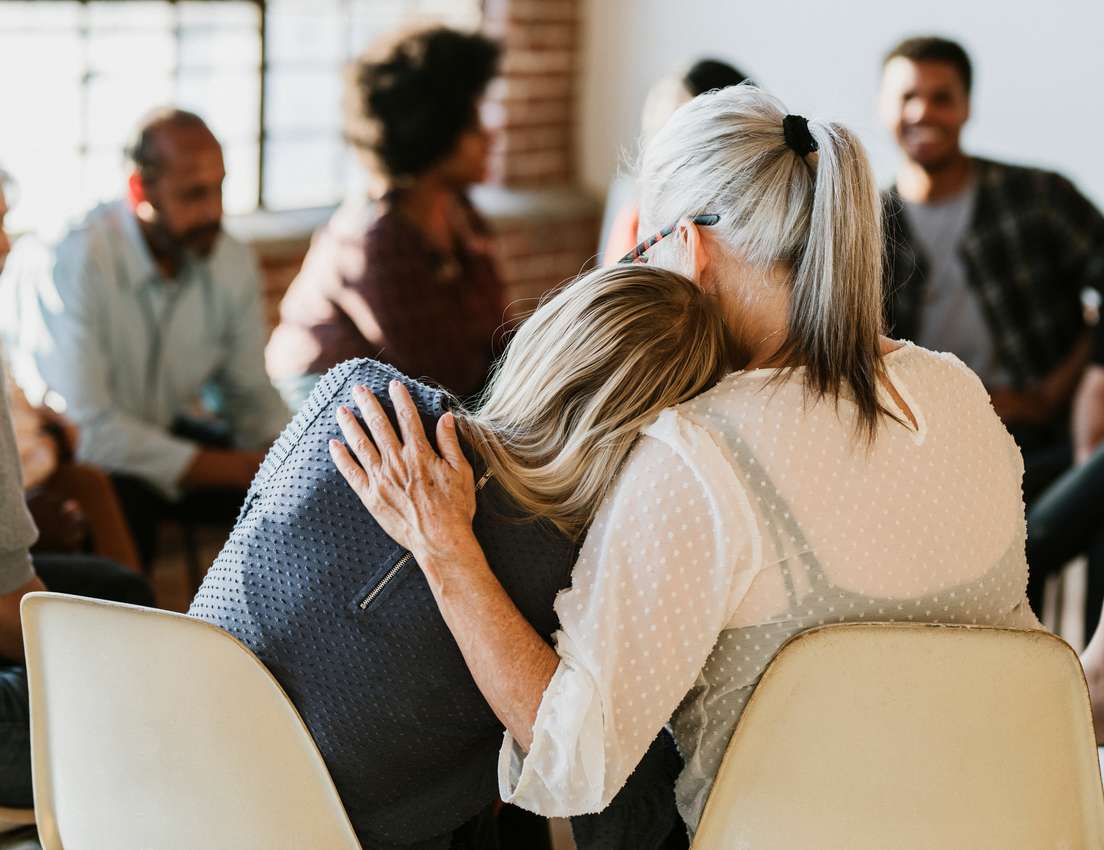 Two women, seen from behind, embrace in a group setting. A grey-haired woman comforts a blonde woman as blurred diverse peopl
