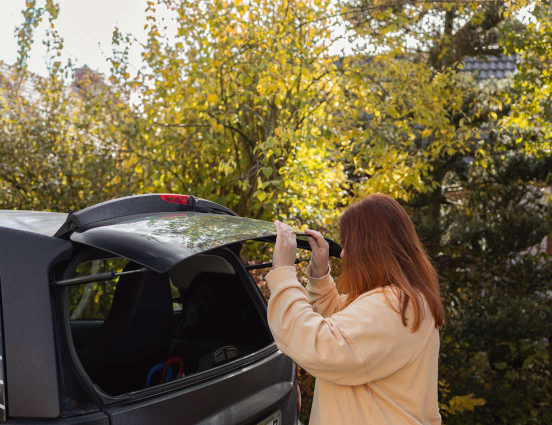 Red-haired woman in peach sweatshirt closing a black car's trunk with a pixelated design. Autumn trees are in the background.