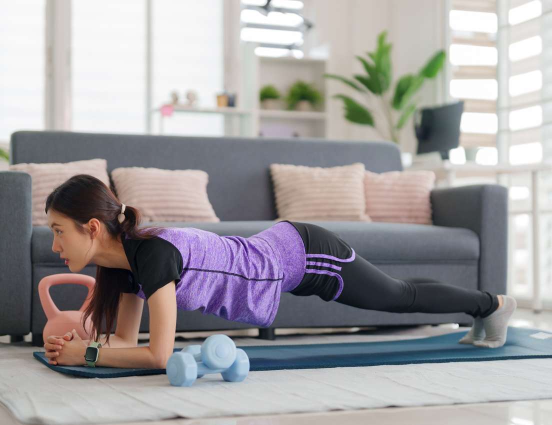 An Asian woman in purple and black activewear holds a forearm plank on a blue yoga mat in a bright living room, with dumbbell