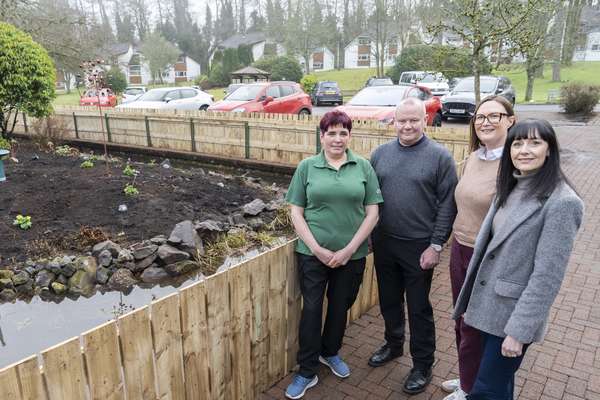 Four people stand beside a newly fenced garden with a pond, dark soil, and small plants. Houses and cars are visible in the background.