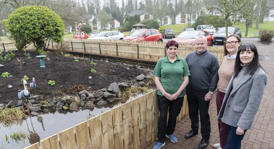 Four people stand beside a newly fenced garden with a pond, dark soil, and small plants. Houses and cars are visible in the b