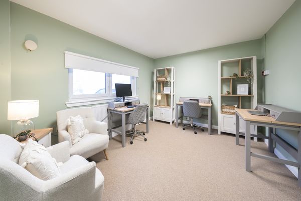 A bright room with light green walls and carpet, two beige armchairs, three grey and wood desks with chairs, and two tall she