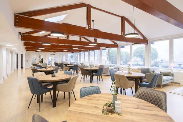 A bright, modern communal dining and lounge area with numerous round wooden tables, comfortable chairs, sofas by large window