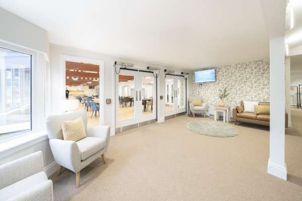 Bright lobby with cream armchairs, a brown leather sofa, and a leaf-patterned wall. Glass doors open to a dining room, a wind
