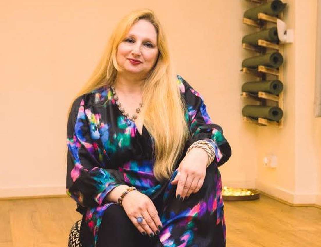 Smiling woman with long blonde hair in a colorful top, seated in a yoga studio with mats on a wall rack.