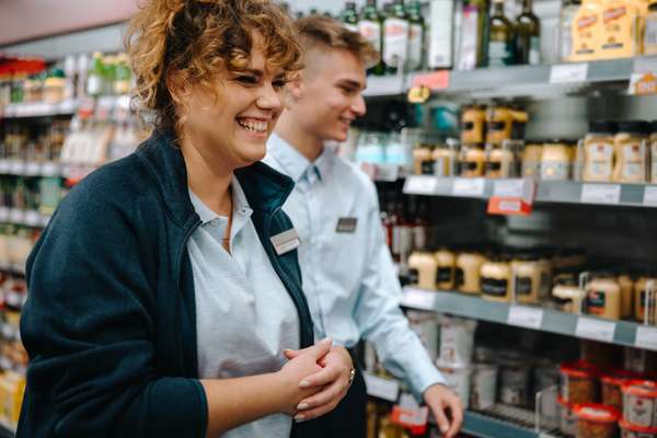 A happy female grocery store worker with curly hair smiles brightly in an aisle, a male colleague and blurred product shelves behind her.