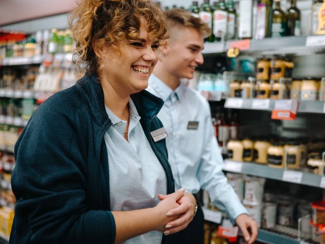 A happy female grocery store worker with curly hair smiles brightly in an aisle, a male colleague and blurred product shelves behind her.