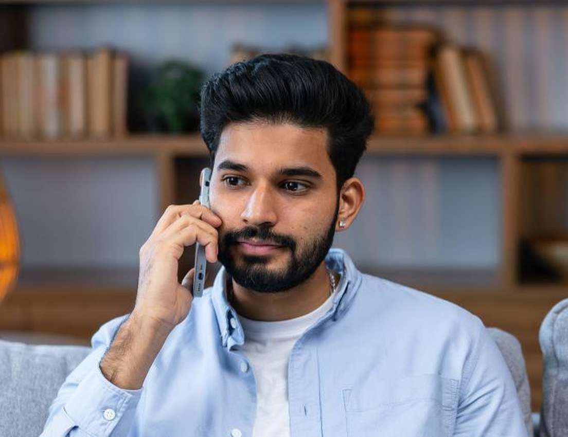 Young bearded man in a light blue shirt talking on a smartphone, sitting on a gray couch with a bookshelf behind him.