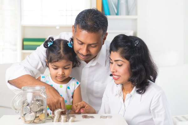 A family of three counts coins together at a table, with a jar of coins beside them, smiling and focused on the task.