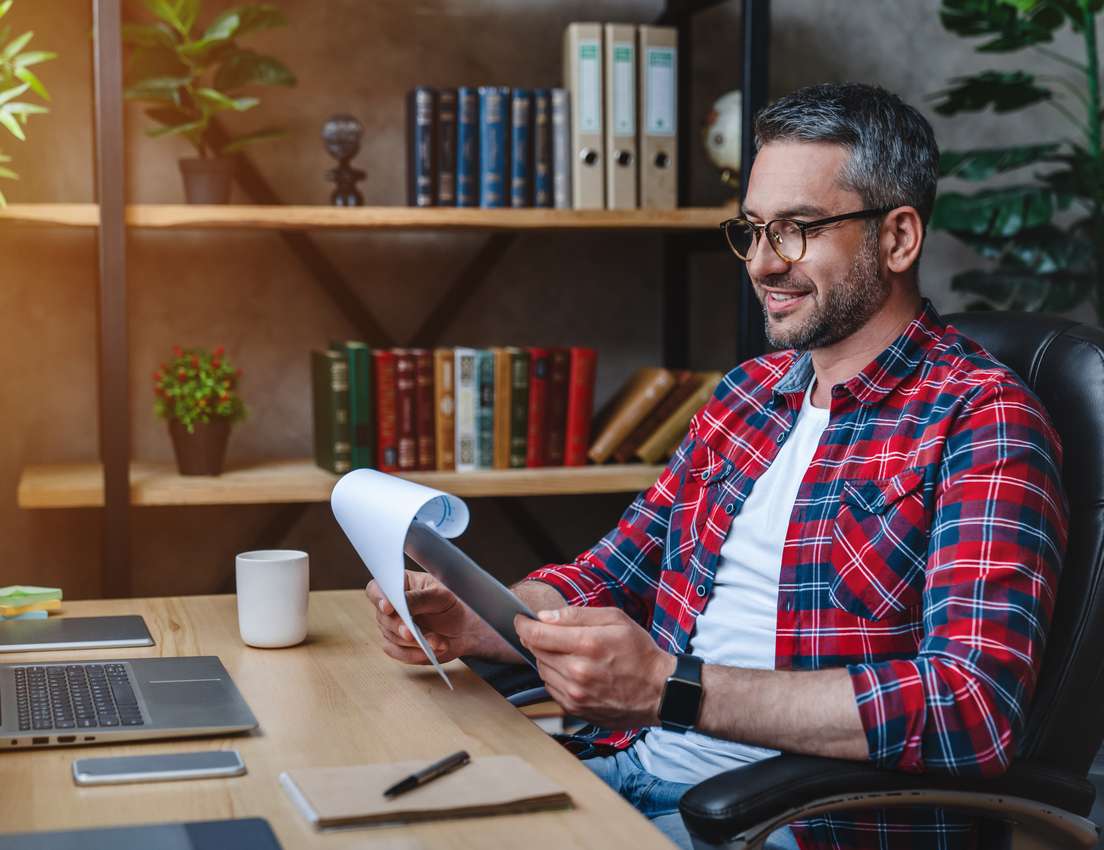 A smiling man in a plaid shirt and glasses sits at a wooden desk, reviewing papers. A laptop, coffee mug, and shelves with bo