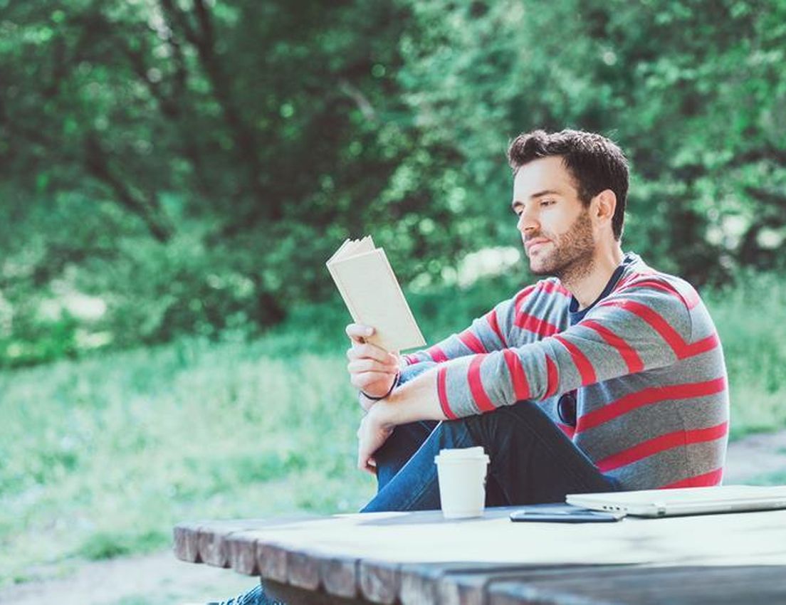 Bearded man in striped shirt reading a book at a picnic table with coffee and laptop in a park.