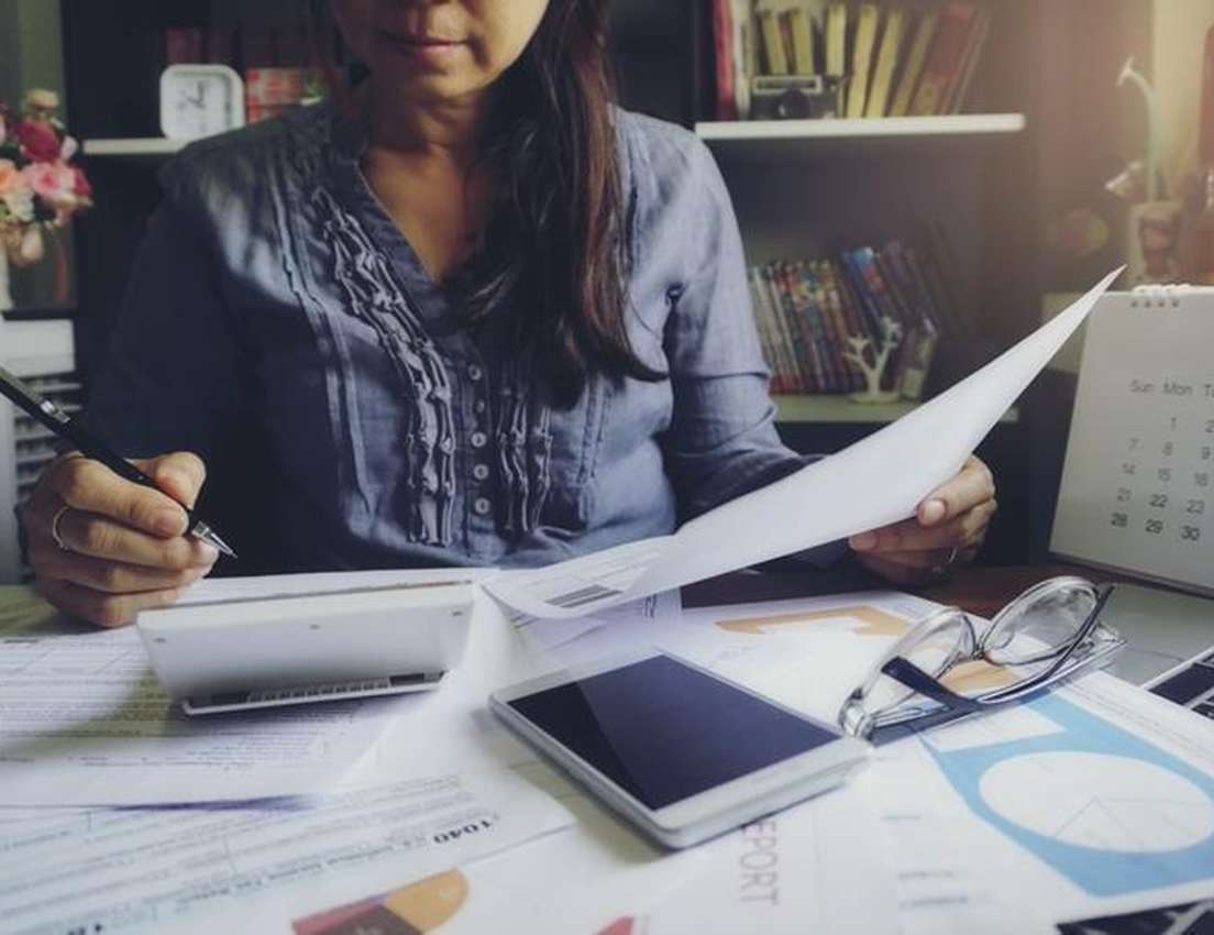 Person calculating taxes with a calculator, pen, and various financial documents including a 1040 form on a cluttered desk.