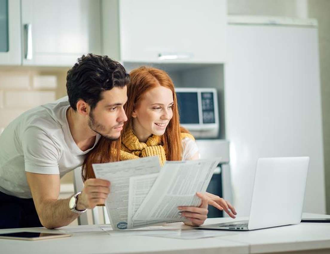 A young couple in a bright kitchen reviewing bills and documents, using a laptop for financial planning.