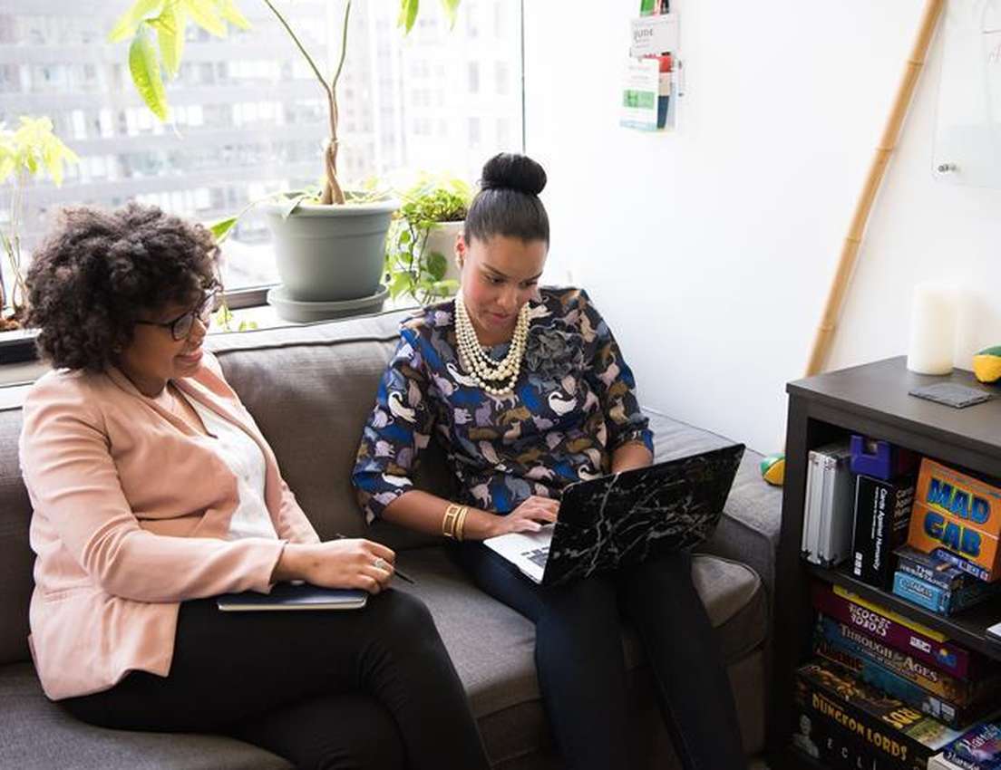 Two women collaborate on a couch in a bright office. One types on a laptop; the other holds a notebook. Plants and bookshelf 
