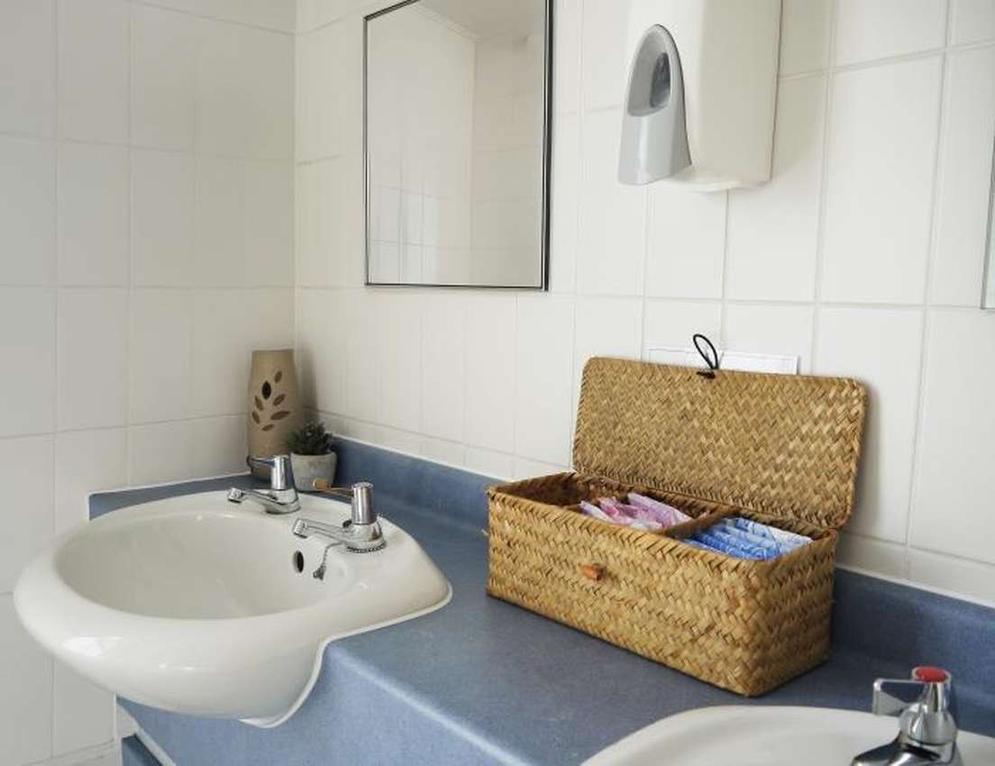Clean public bathroom with two white sinks, a mirror, and a straw basket filled with menstrual products on a blue counter.