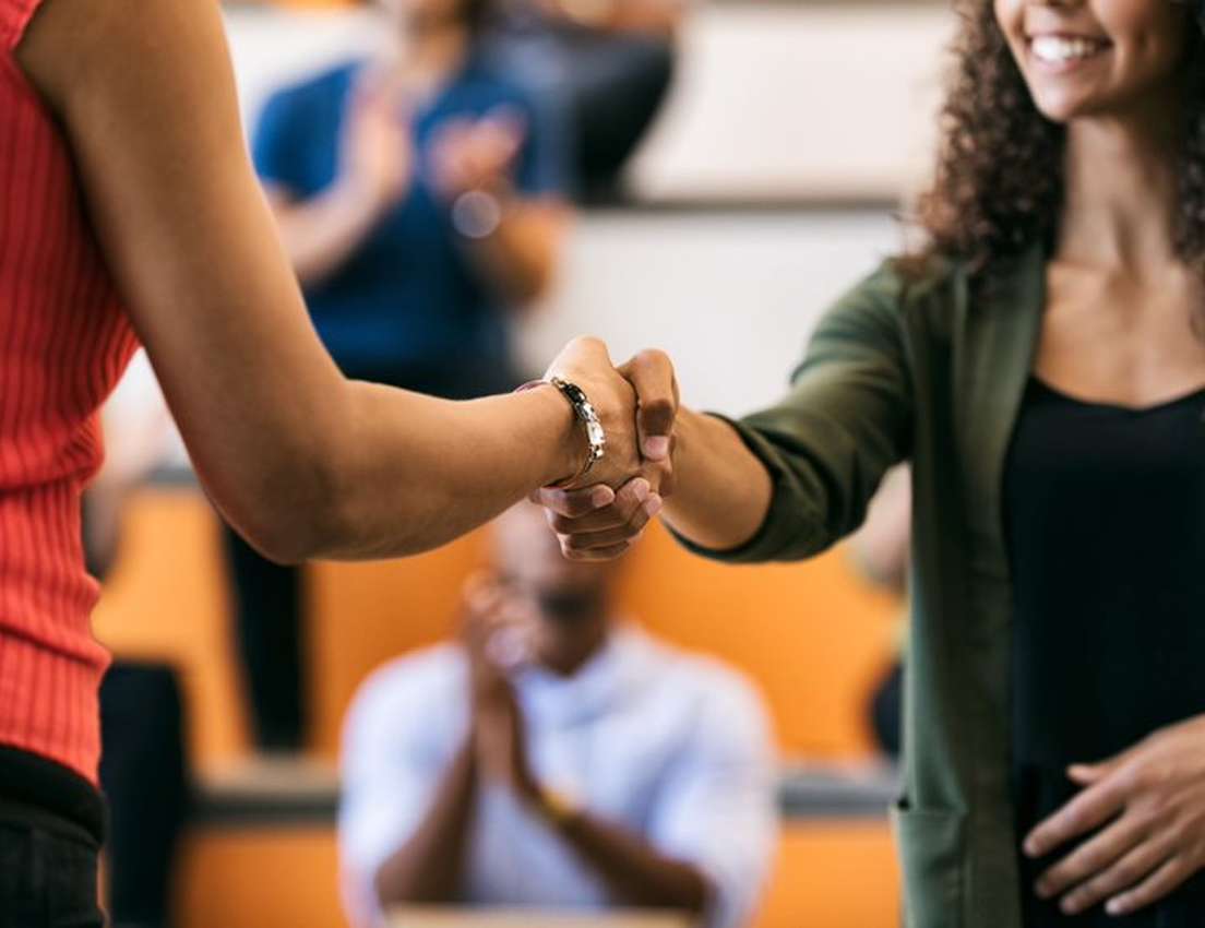 Two women shaking hands, one in red, the other in green with a smiling face. Blurred people in the background are clapping.