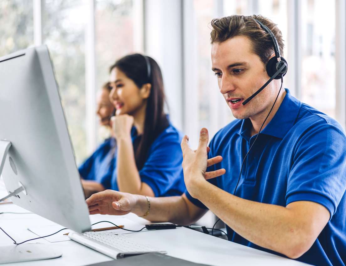 A man in a blue polo shirt and headset actively speaks at a computer, gesturing. Other call center agents work in the backgro