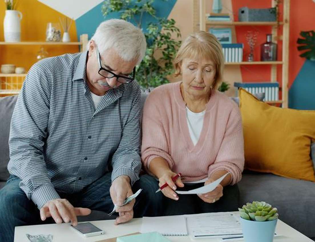An older couple sits on a couch, man looking at phone and cash, woman holding documents and a pen, appearing to manage financ