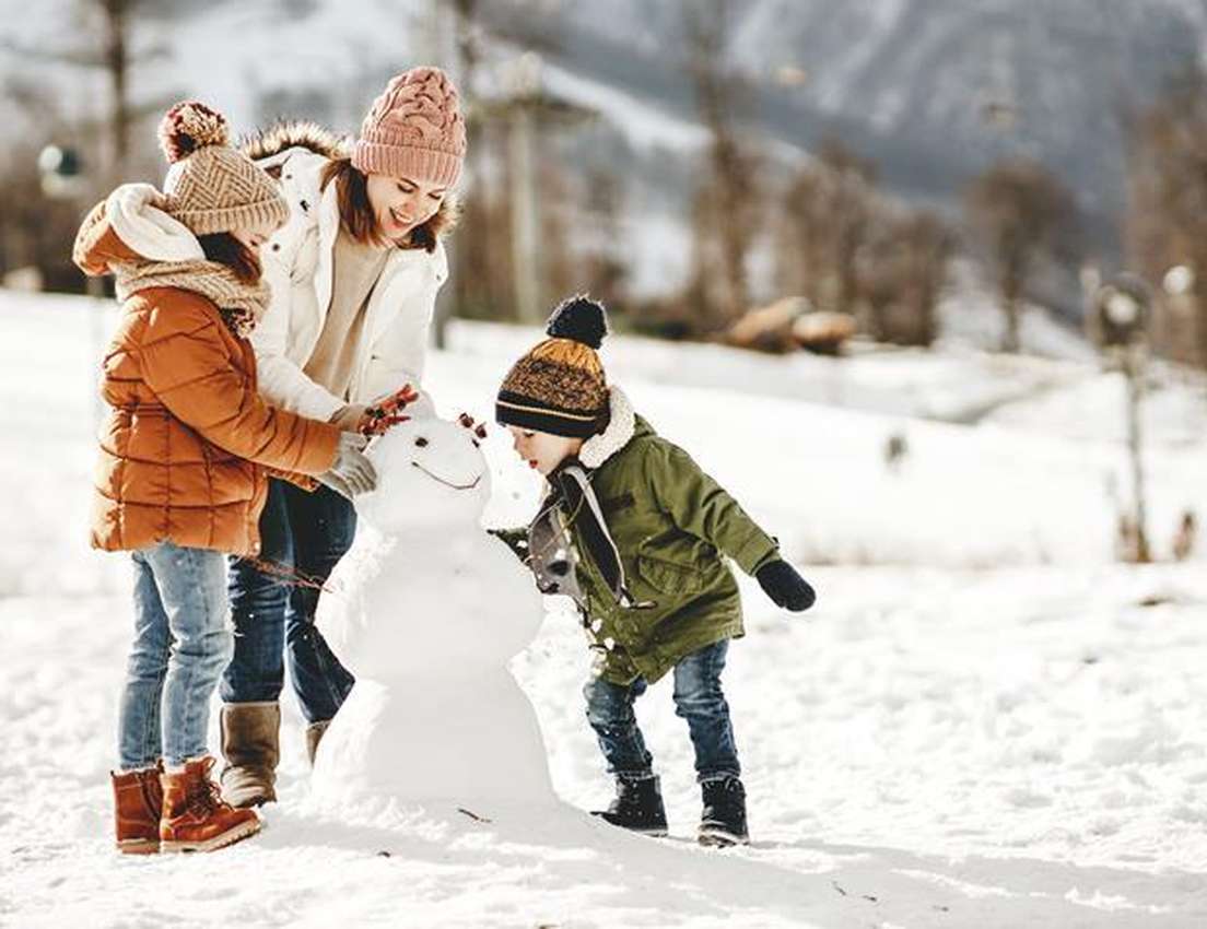 A mother and two children bundled in winter clothes happily building a snowman together in a sunny, snowy landscape.