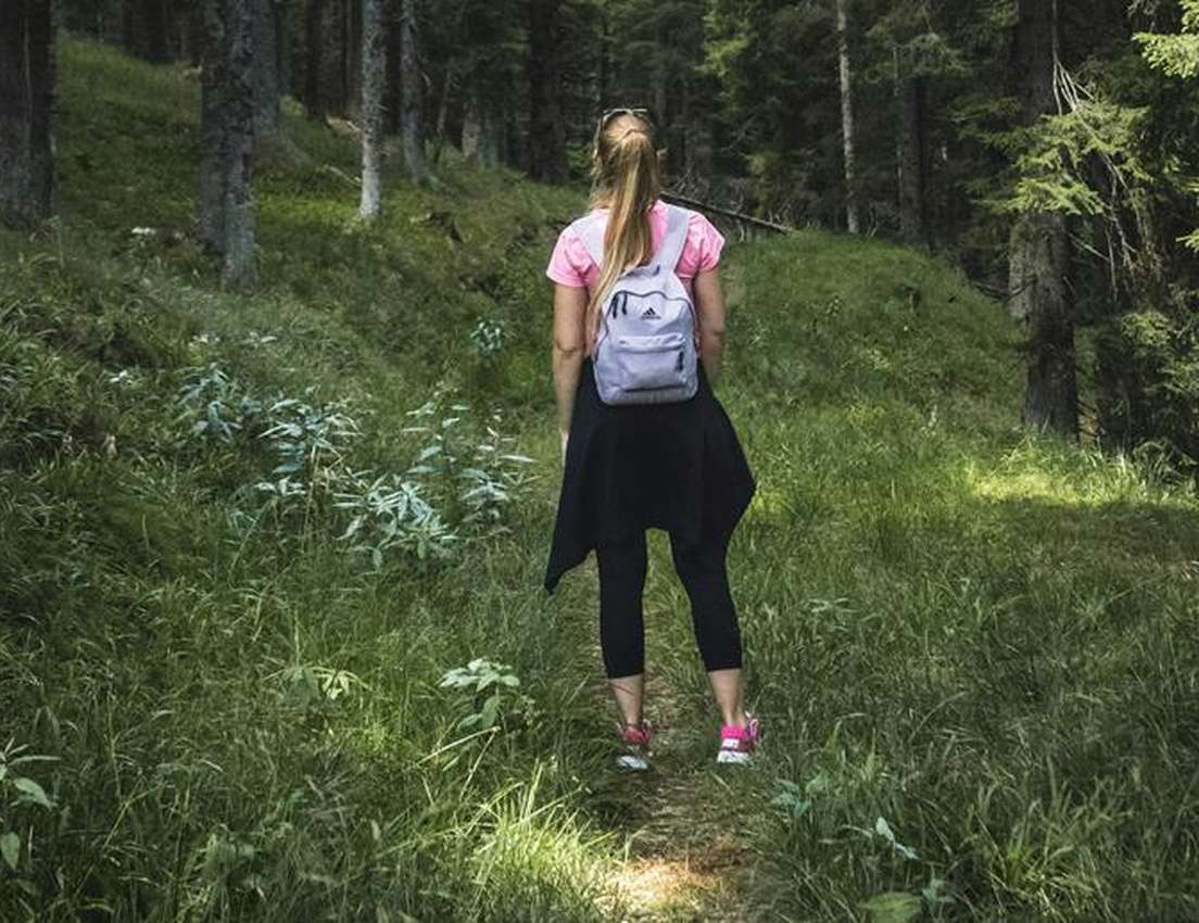 Woman with a ponytail and grey backpack hiking a grassy path through a lush, green forest on a sunny day.