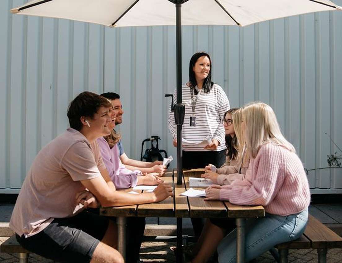 An instructor leads a group of young adults at an outdoor picnic table under a white umbrella. They have papers and appear en