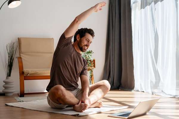 A man with a beard sits on a mat, stretching his arm overhead while looking at a laptop in a bright room with a window.