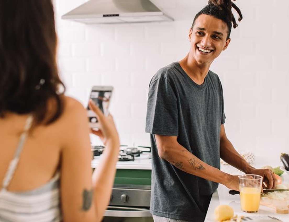 Woman photographs a smiling man with dreadlocks and nose ring chopping vegetables in a bright kitchen with fresh produce.