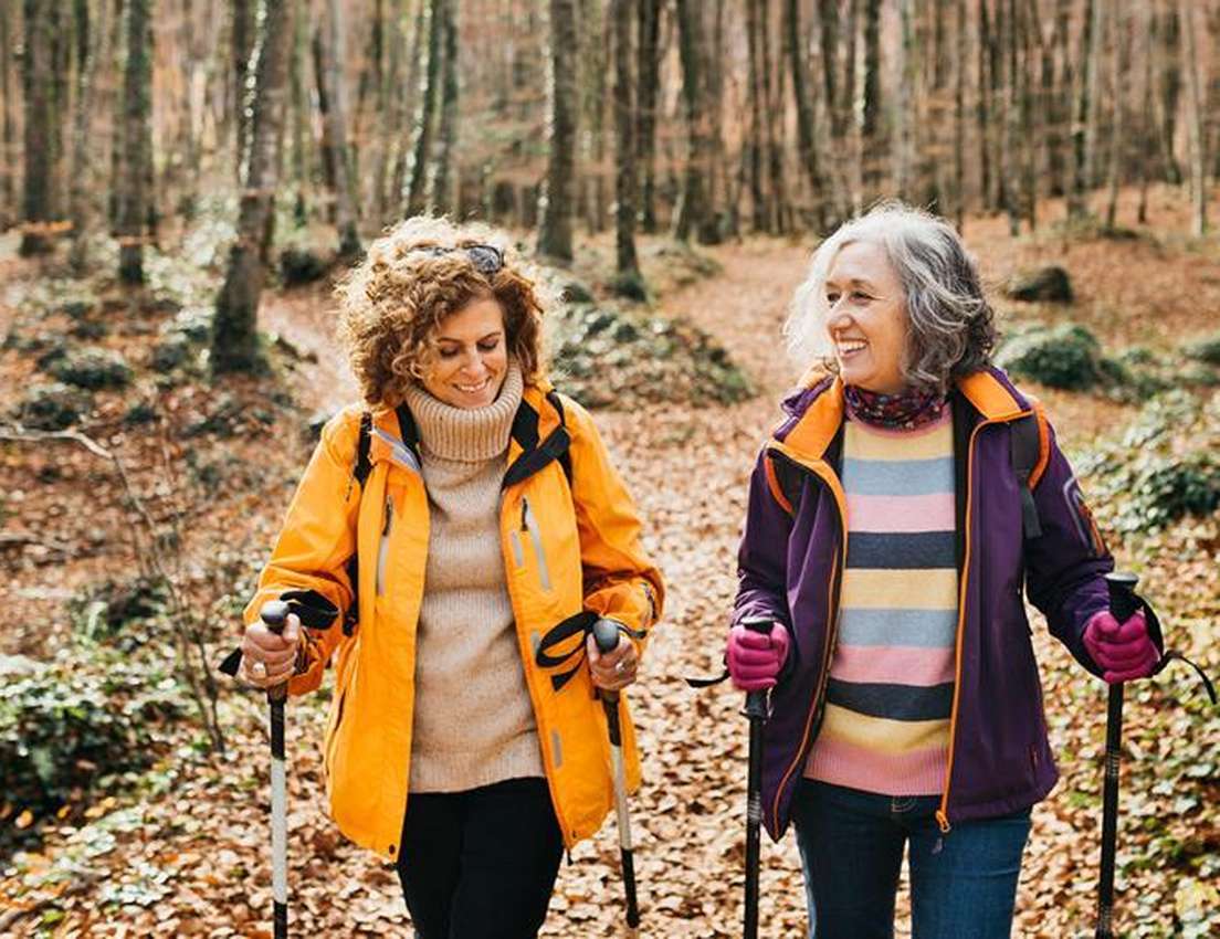 Two smiling women hike with poles through an autumn forest, leaves covering the ground. One wears an orange jacket, the other