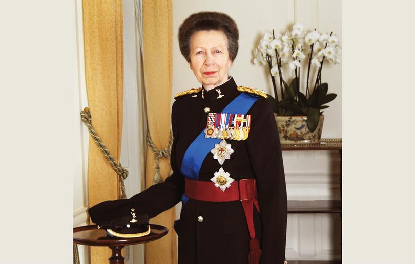 Princess Anne in full military uniform with medals, a blue sash, and a red sash, standing next to a table with a cap. White orchids in background.