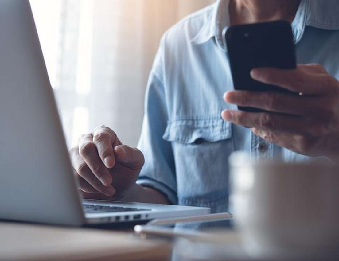 A person in a blue shirt is actively using a laptop while holding a smartphone. A coffee cup and stylus are on the desk.