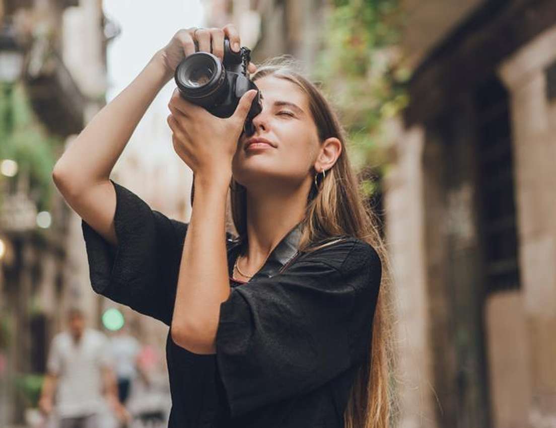 Young woman with closed eyes taking a photo with a DSLR camera on an old European street.
