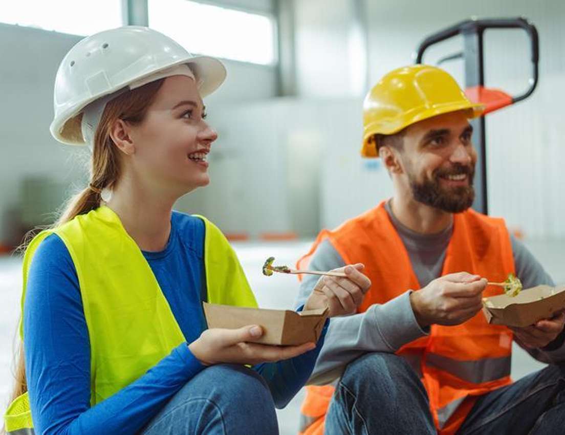 A smiling woman and man in hard hats and safety vests eat lunch from cardboard containers in a warehouse.