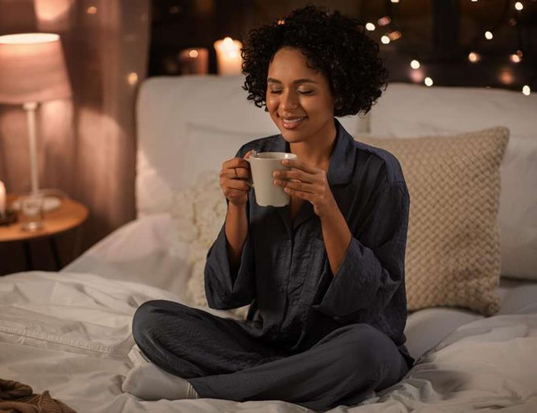 A smiling woman with curly hair, wearing pajamas, sits cross-legged on a bed, holding a mug, in a cozy, dimly lit bedroom.