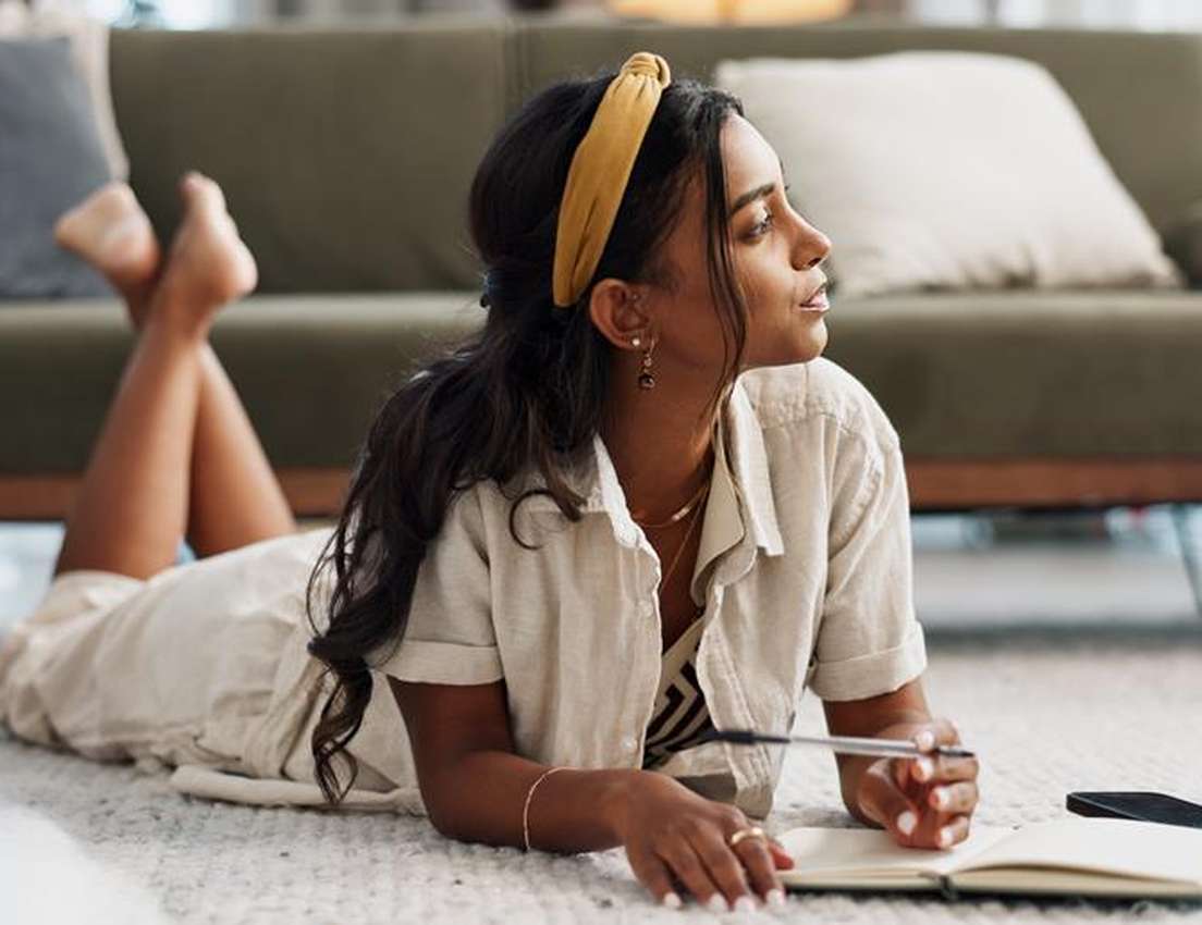 A young woman with a yellow headband lies on a rug, holding a pen over a notebook, looking thoughtfully to the side.