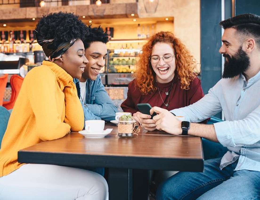 Four diverse friends laugh heartily while looking at a smartphone together at a cafe table.