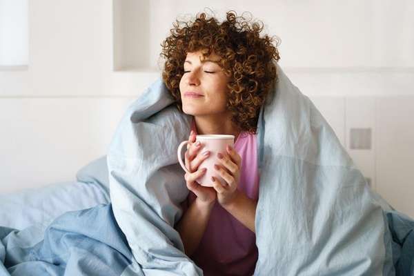A woman with curly hair wrapped in a blue duvet in bed, holding a pink mug with her eyes closed, looking content.