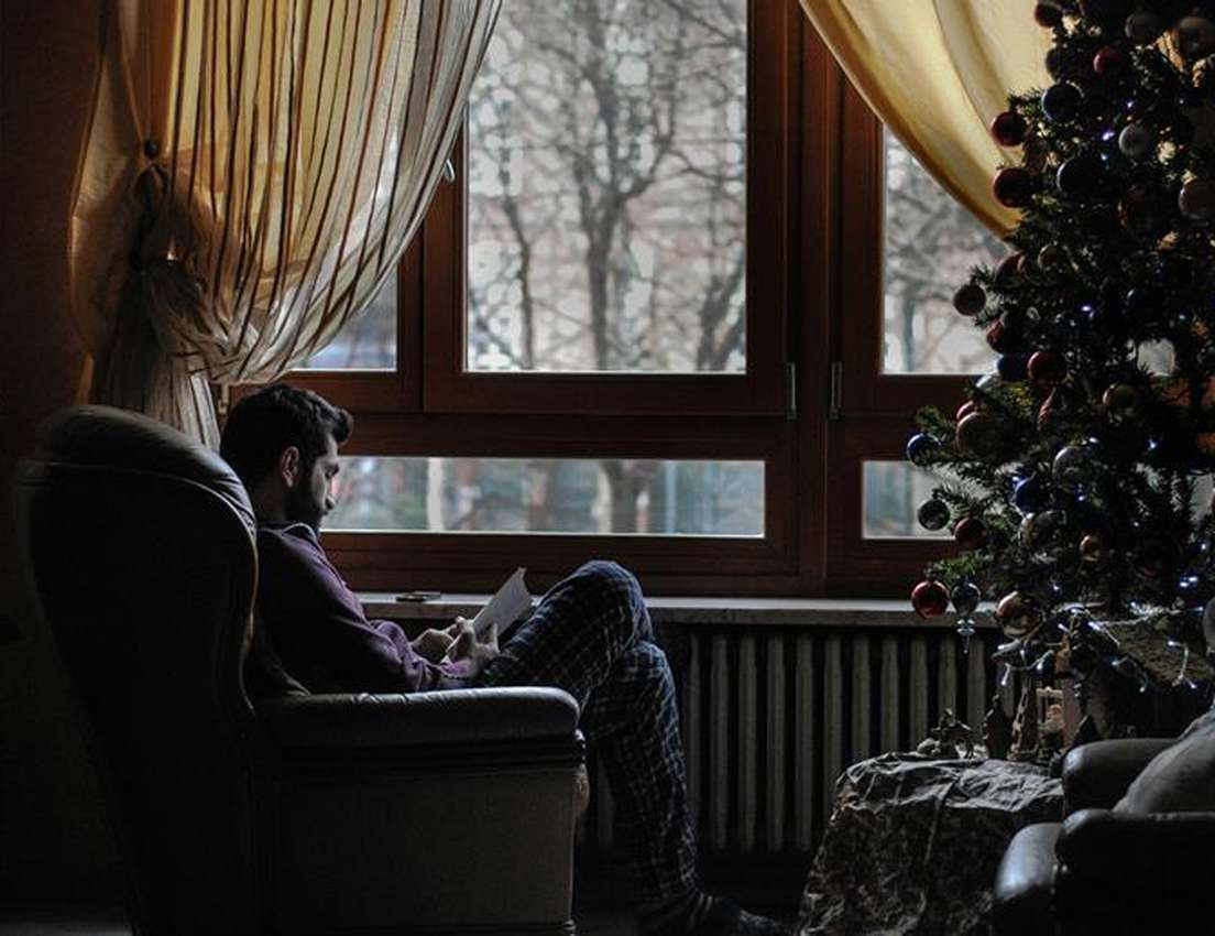 Man reads in an armchair by a large window with bare trees outside, next to a lit Christmas tree. Cozy winter holiday scene.