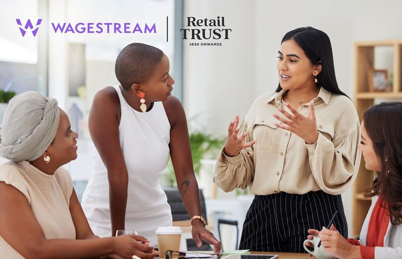 Four diverse women in a collaborative office meeting, with one speaking expressively to the others around a table. Logos visible.