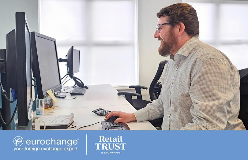 A happy bearded man in glasses smiles while using a computer mouse at an office desk with multiple screens.