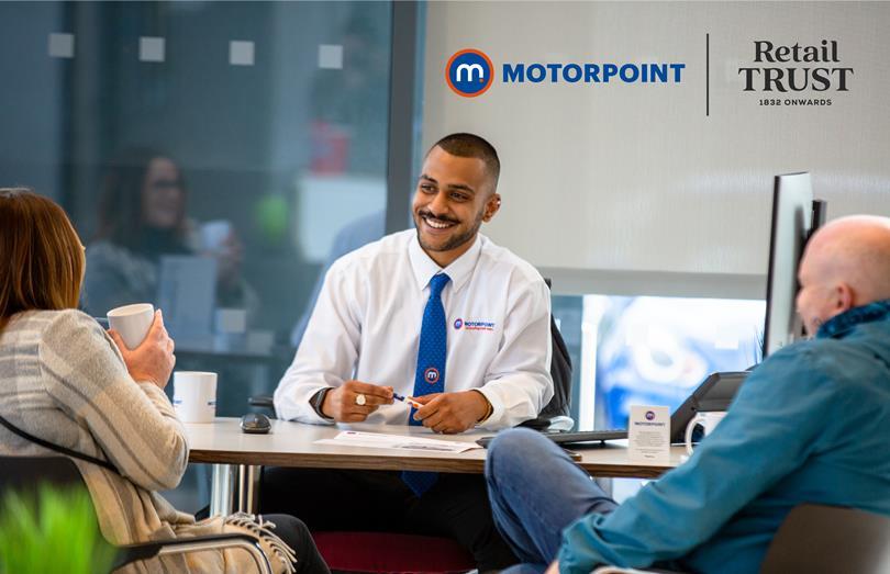 Smiling Motorpoint employee in a white shirt and blue tie talking to two customers at a desk. Motorpoint and Retail Trust logos visible.