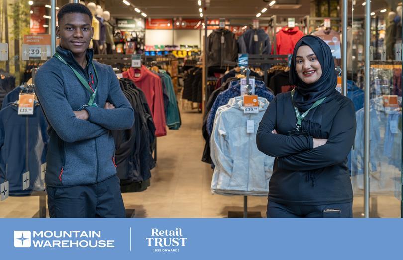 Two smiling retail employees, a man and a woman in a hijab, stand proudly in a Mountain Warehouse store with clothing racks behind them.