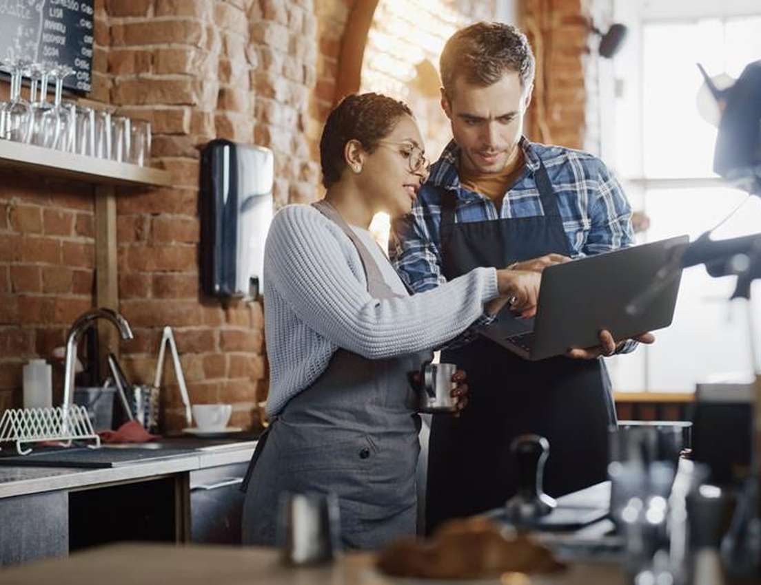 Two cafe owners, a man and a woman, collaborate using a laptop in their rustic coffee shop with brick walls and espresso mach
