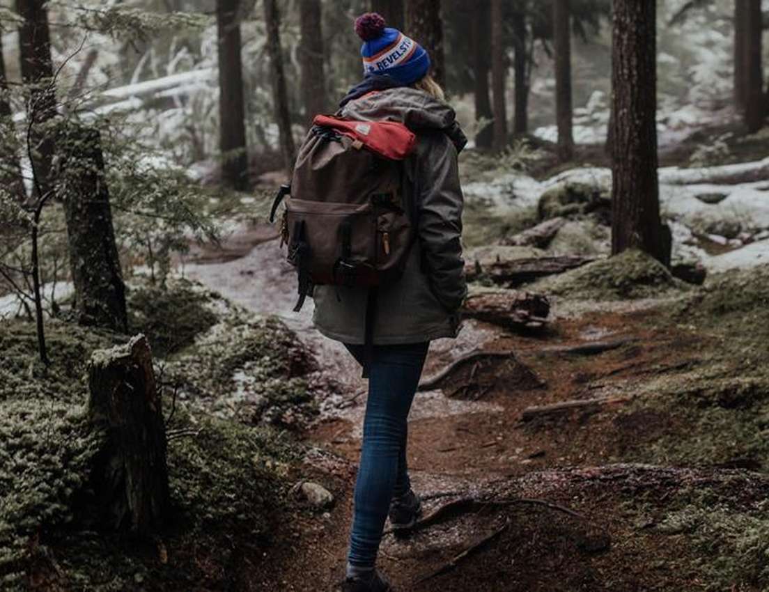 A person with a backpack and beanie walks away on a snowy, mossy forest path.