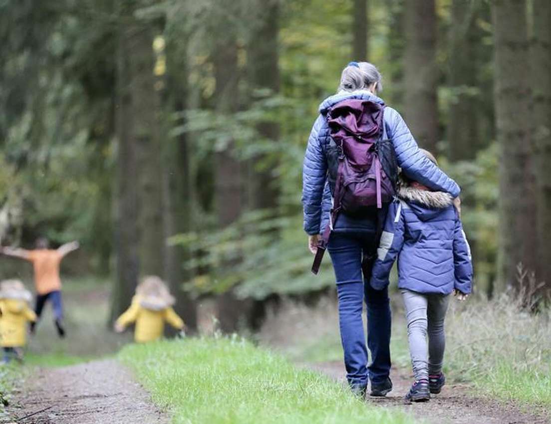 A woman and child, wearing coats, walk along a forest path. Other children run ahead in yellow jackets.