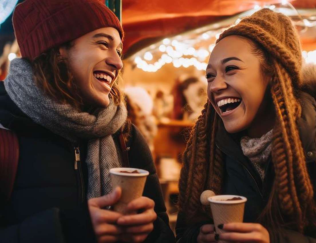 Two young people in winter clothes laugh and hold hot drinks at a festive outdoor market with warm lights.
