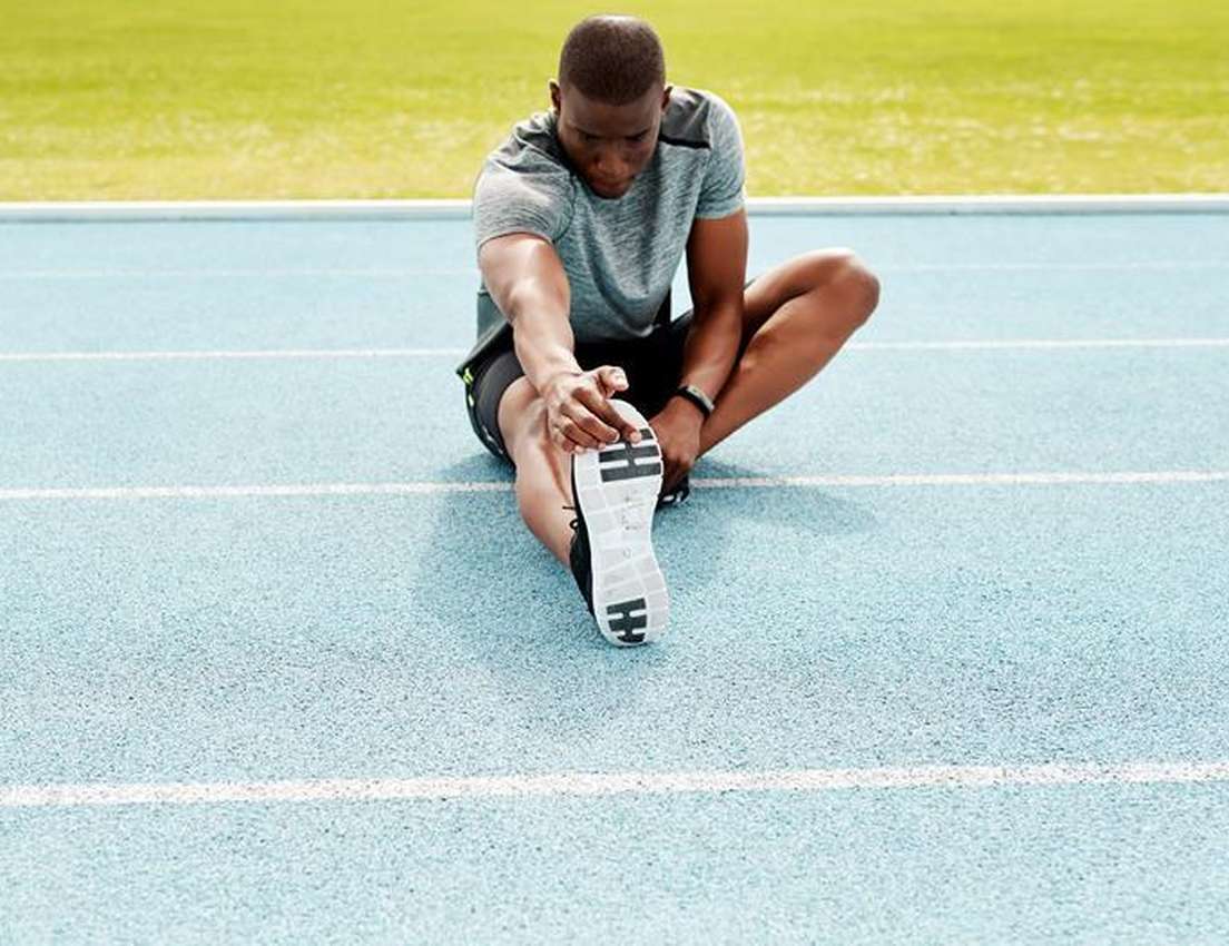 A man in athletic wear sits on a blue running track, stretching one leg and reaching for his foot.