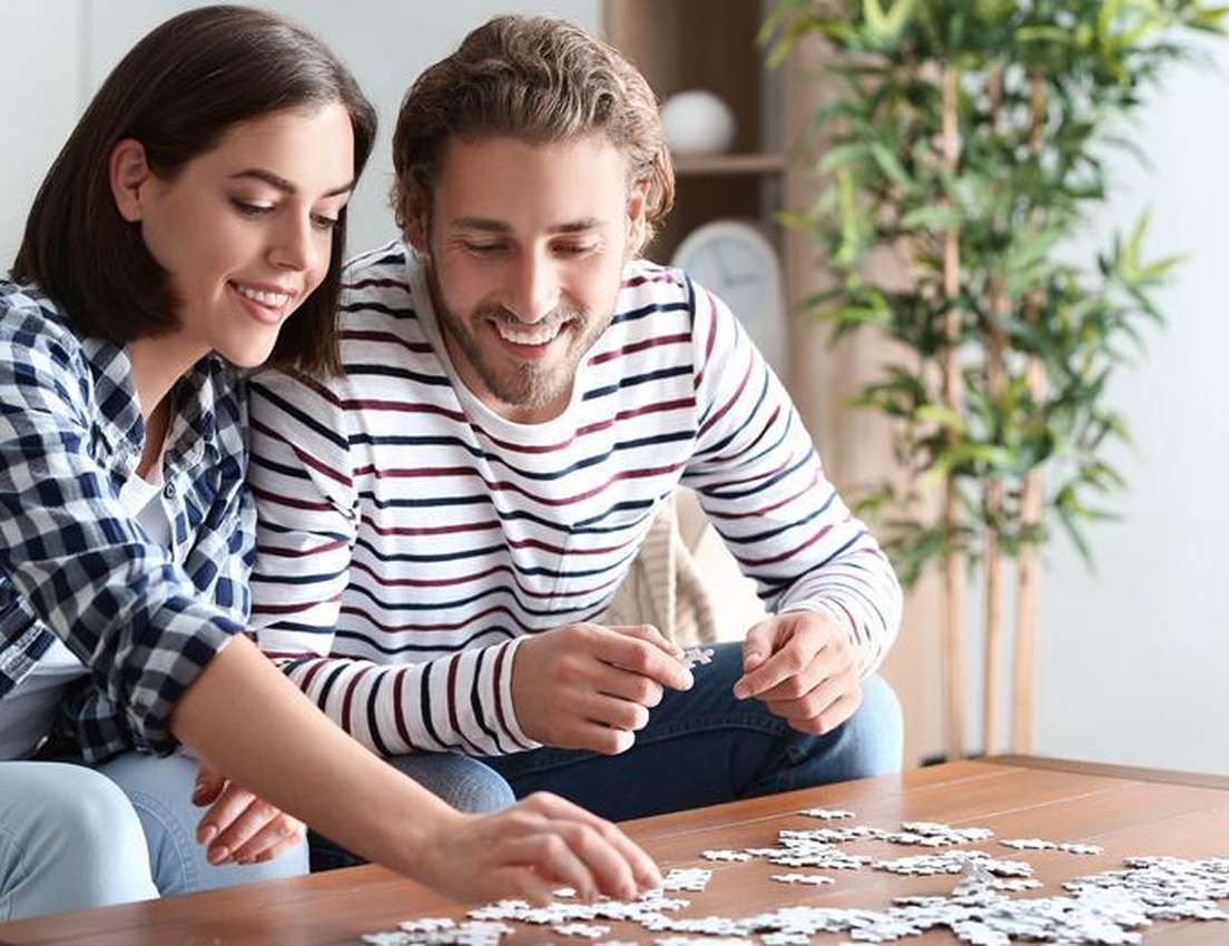 A smiling man and woman focus on assembling a jigsaw puzzle together on a wooden coffee table in a bright living room.