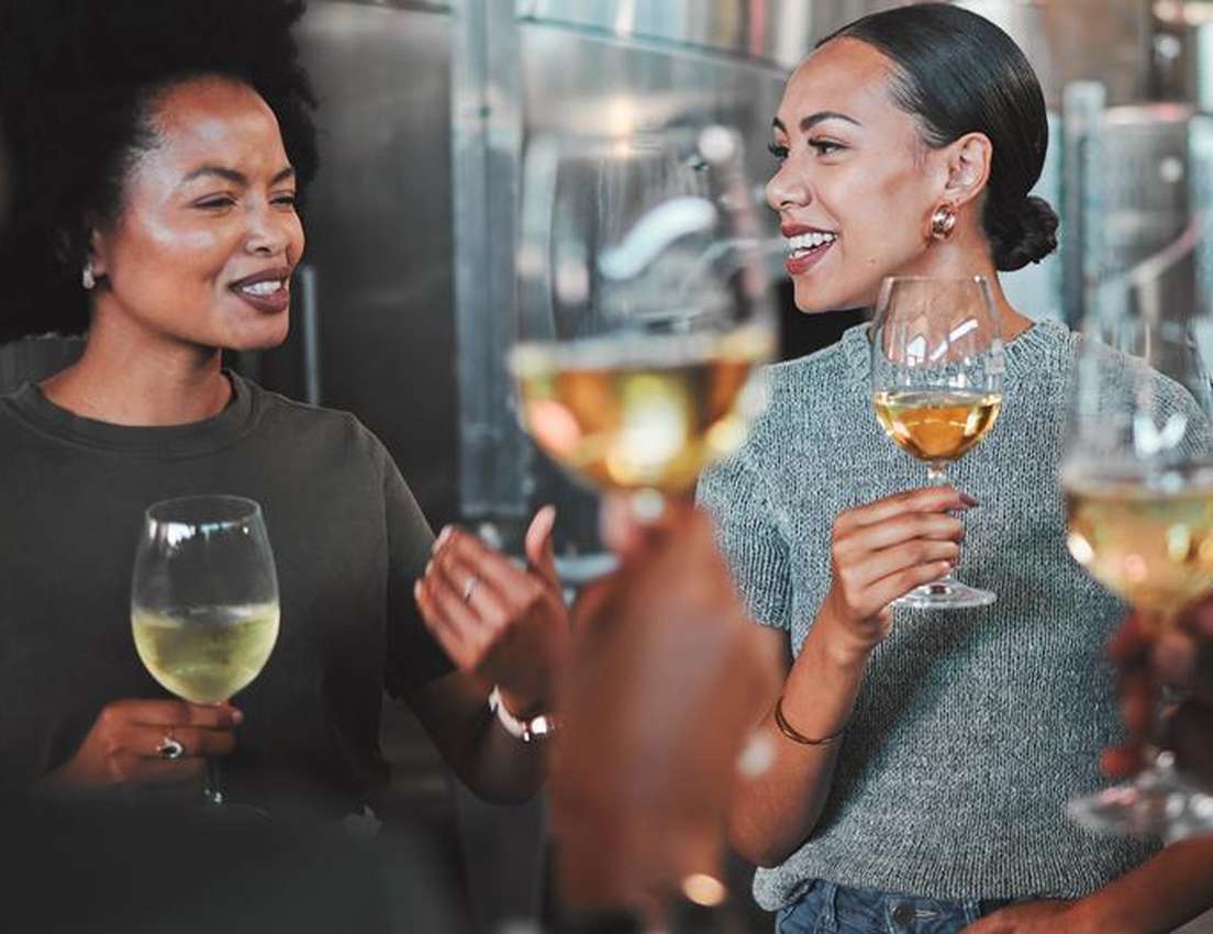 Two smiling women tasting white wine in a winery, engaged in conversation with friends.